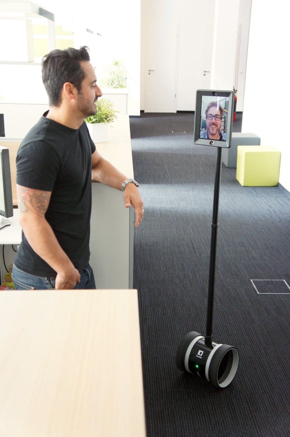 Netzkern-Mitarbeiter Taylan Demirkol (links) spricht mit seinem Hamburger Kollegen Christopher Loos (Bildschirm) über den Telepräsenzroboter Double. Letzerer kann sich dank des Roboters frei in der Wuppertaler Agentur bewegen und die Kollegen selbständig aufsuchen, mit denen er etwas zu besprechen hat.
