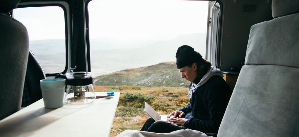 Ein junger Mann sitzt in einem Van in der Natur, einen Laptop auf dem Schoß und eine Mütze auf dem Kopf. Eine Kaffeekanne und -becher im Vordergrund auf dem Tisch im Auto.