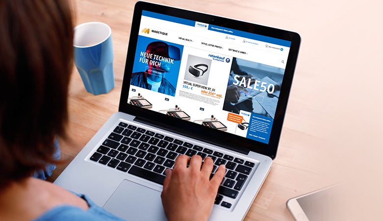 Man schaut über die Schulter einer Frau auf ihren Laptop. Das Bild zeigt einen generischen Onlineshop mit einem großen Sale. Links neben dem Laptop steht eine leere Kaffeetasse.