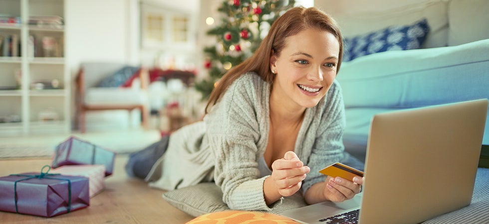 Eine Frau shoppt an Wihnachten im Internet. Sie liegt auf dem Wohnzimmerboden mit einer goldenen Kreditkarte in der Hand vor dem Laptop. Im Hintergrund sieht man einen Weihnachtsbaum und einige Geschenke.