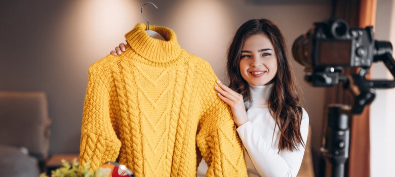 Auf dem Bild sieht man eine Frau mit braunen Haaren, die einen gelben Pullover hochhält. Im Vordergrund sieht man unscharf eine Kamera, die die Frau filmt.
