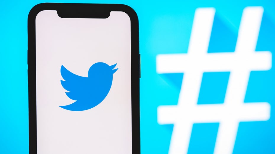 Smartphone mit angezeigtem Twitter-Logo vor einem blauen Hintergrund mit weißem Hashtag