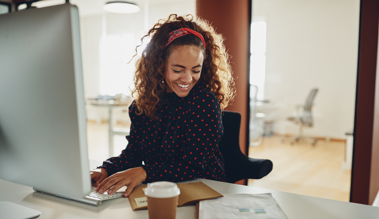 Eine junge Frau lachend am Schreibtisch, vor ihr ein iMac sowie Dokumente und ein Kaffee zum Mitnehmen. Im Hintergrund helle Büroräume