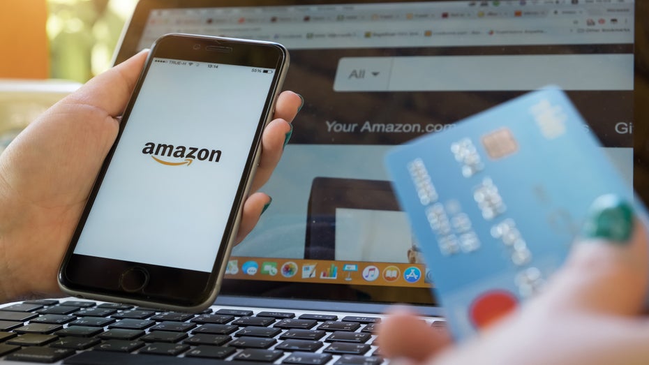 Eine Frau sitzt mit einer Kreditkarte in der Hand vor einem Computer, auf dessen Bildschirm die Amazon-Website zu sehen ist. (Foto: Shutterstock.com / Worawee Meepian)