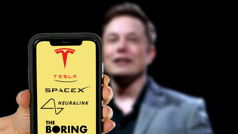 Elon Musk – im Vordergrund ist ein Smartphone mit den Logos seiner Unternehmen zu sehen. (Bild: Shutterstock.com/Kavi Designs)
