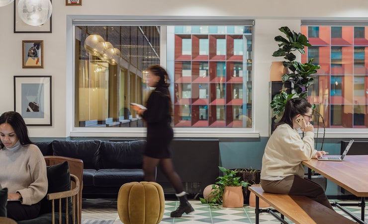 In einem schön gestalteten Bürogebäude, das eher an eine bar erinnert, arbeiten zwei Frauen an zwei verschiedenen Tischen. Eine dritte Frau läuft von rechts nach links.