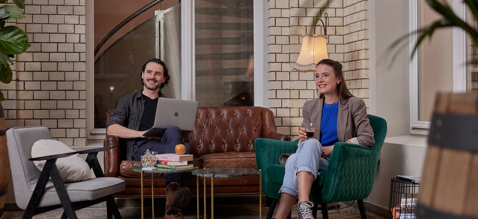 Zwei Menschen sitzen in einer Sofa und Couchecke. Der Mann arbeitet an seinem MacBook, die Frau trinkt einen Wein. 