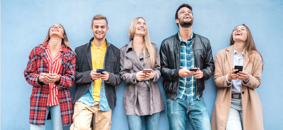 Eine weiße Freundesgruppe guckt nach oben, während sie alle ein Handy in der Hand halten. Der Hintergrund ist blau.