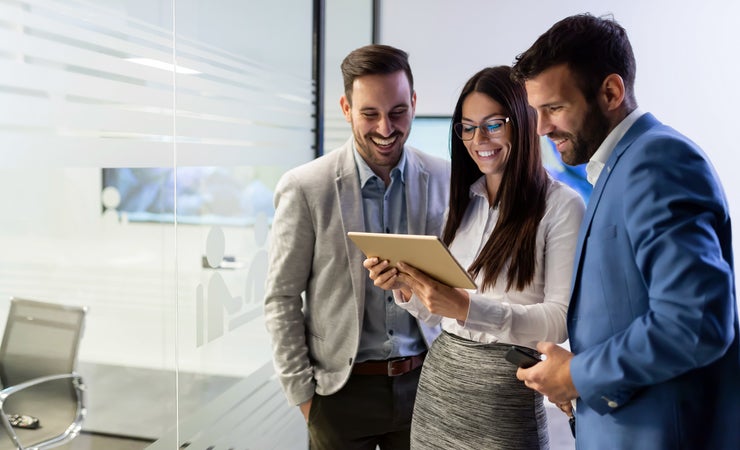 Zwei Männer im Anzug schauen einer Frau im Business-Look auf ihr Tablet. Alle lächeln.