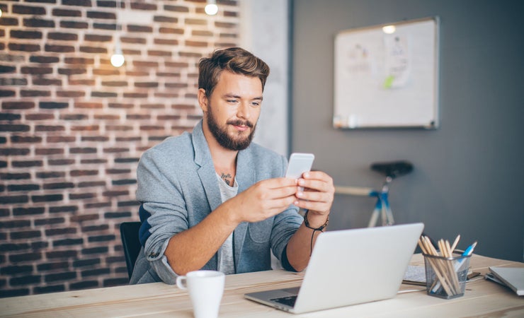 Ein weißer Mann mit Vollbart sitzt im Büro. Hinter ihm ist eine industriell aussehende Ziegelwand. Auf seinem Schreibtisch steht ein Laptop und ein Kaffee. Der Mann hat die Ärmel seines grauen Jacketts hochgekrämpelt und hat sein Handy in der Hand.