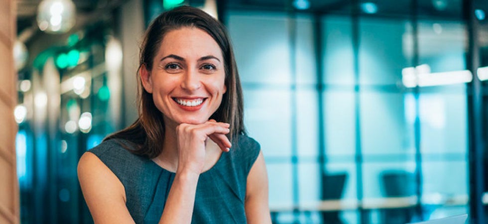 Eine weiße Frau grinst in die Kamera. Sie ist in einem Office-Space und ist business-casual gekleidet.