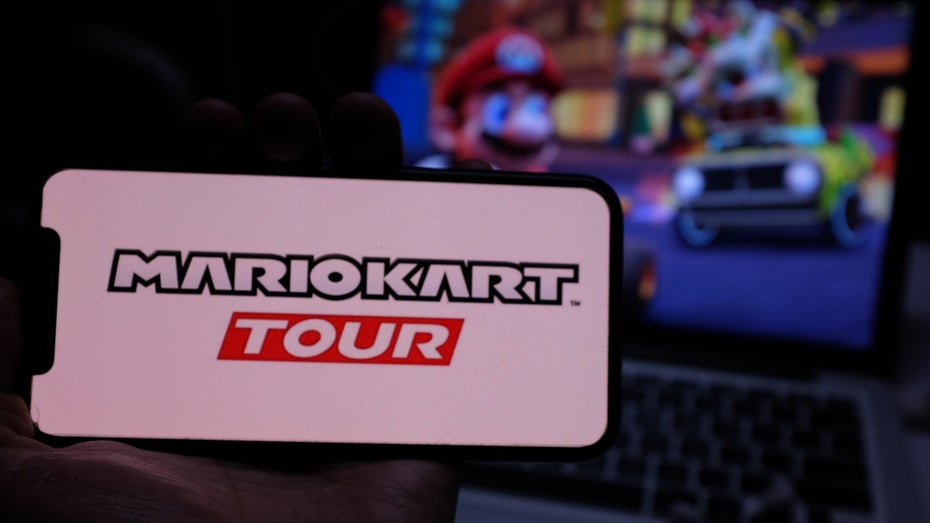 Smartphone-Display mit dem Logo von Mario Kart Tour, im Hintergrund das Spiel auf einem Bildschirm (Bild: Shutterstock / DANIEL CONSTANTE)